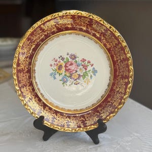 May include: A decorative plate with a floral design. The plate features a cream-colored center with a bouquet of pink, yellow, blue, and purple flowers. The rim is burgundy with a gold floral pattern and a gold edge. The plate is displayed on a black stand.