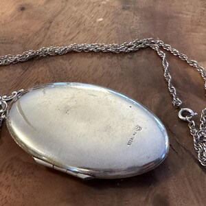Puede incluir: Collar con un medall&oacute;n ovalado de plata de ley y una cadena delicada. El medall&oacute;n cerrado tiene una superficie brillante y reflectante. La palabra "STERLING" est&aacute; estampada en el medall&oacute;n. El collar est&aacute; sobre madera.