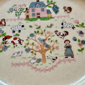 May include: A cream-colored ceramic plate with a farm scene design. The plate features a pink house, farm animals, a tree, and a woman in a dress. The plate has a scalloped edge and is decorated with flowers and a pink fence.