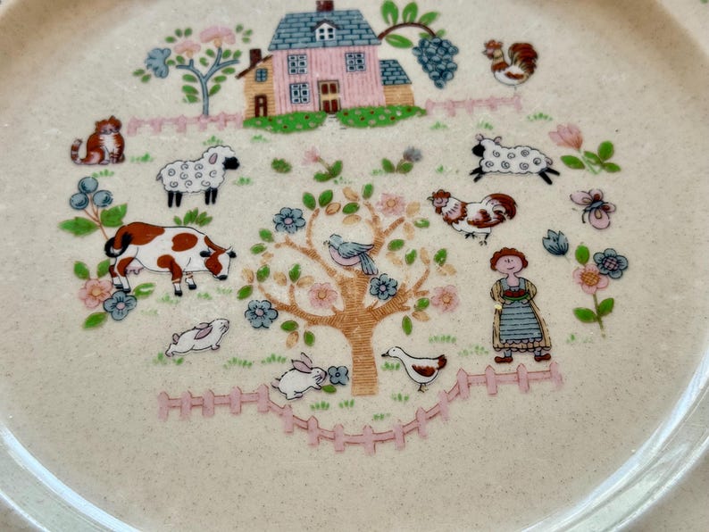 May include: A cream-colored ceramic plate with a farm scene design. The plate features a pink house, farm animals, a tree, and a woman in a dress. The plate has a scalloped edge and is decorated with flowers and a pink fence.