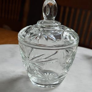 May include: A clear glass sugar bowl with a matching lid featuring a pointed finial. The exterior surface of the bowl and lid is decorated with a pressed starburst pattern, creating a faceted texture that catches the light across the entire vessel.