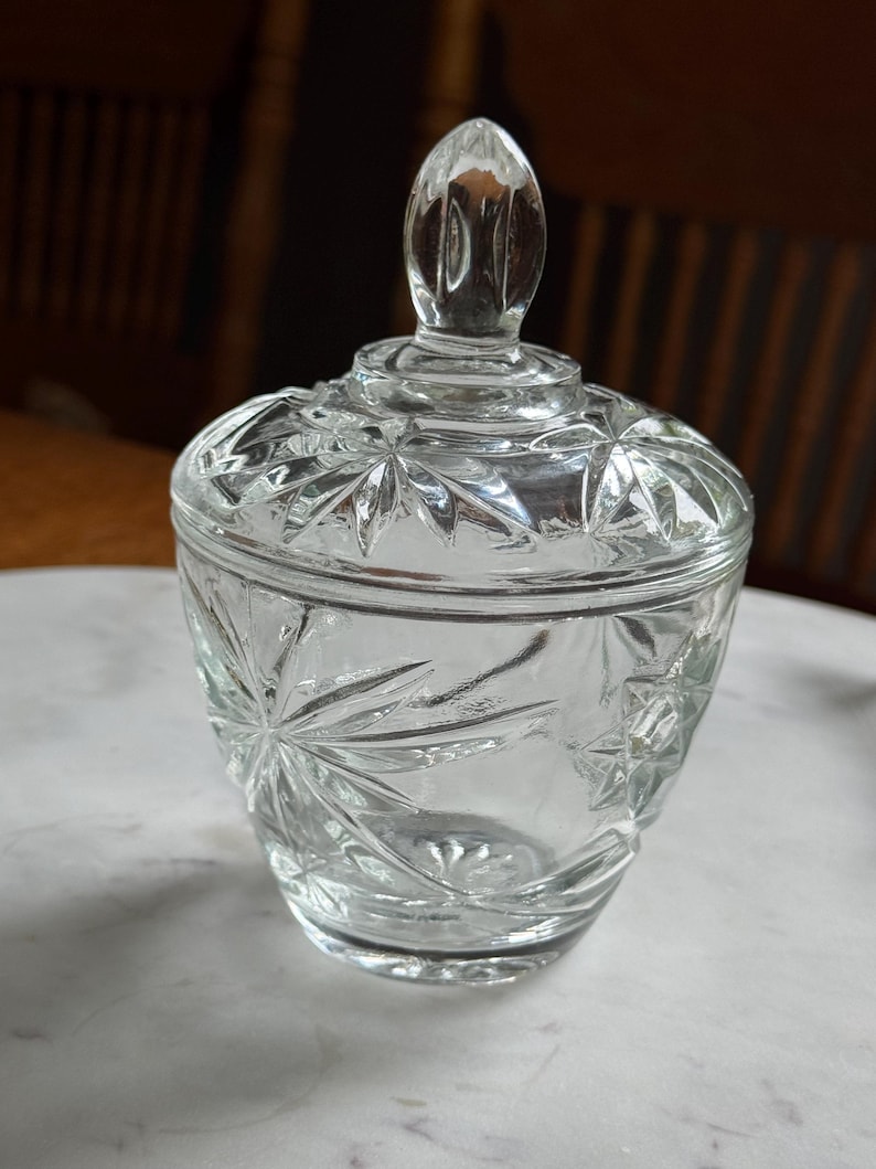 May include: A clear glass sugar bowl with a matching lid featuring a pointed finial. The exterior surface of the bowl and lid is decorated with a pressed starburst pattern, creating a faceted texture that catches the light across the entire vessel.
