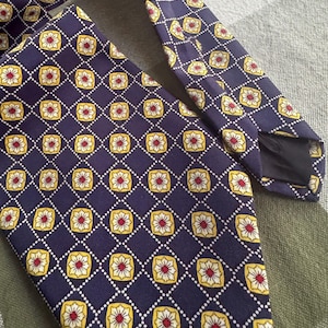 Puede incluir: Una corbata de color morado oscuro con un patr&oacute;n geom&eacute;trico repetitivo de l&iacute;neas de diamantes punteadas en blanco. Dentro de cada diamante hay un cuadrado amarillo de esquinas redondeadas con un motivo floral blanco y rojo.