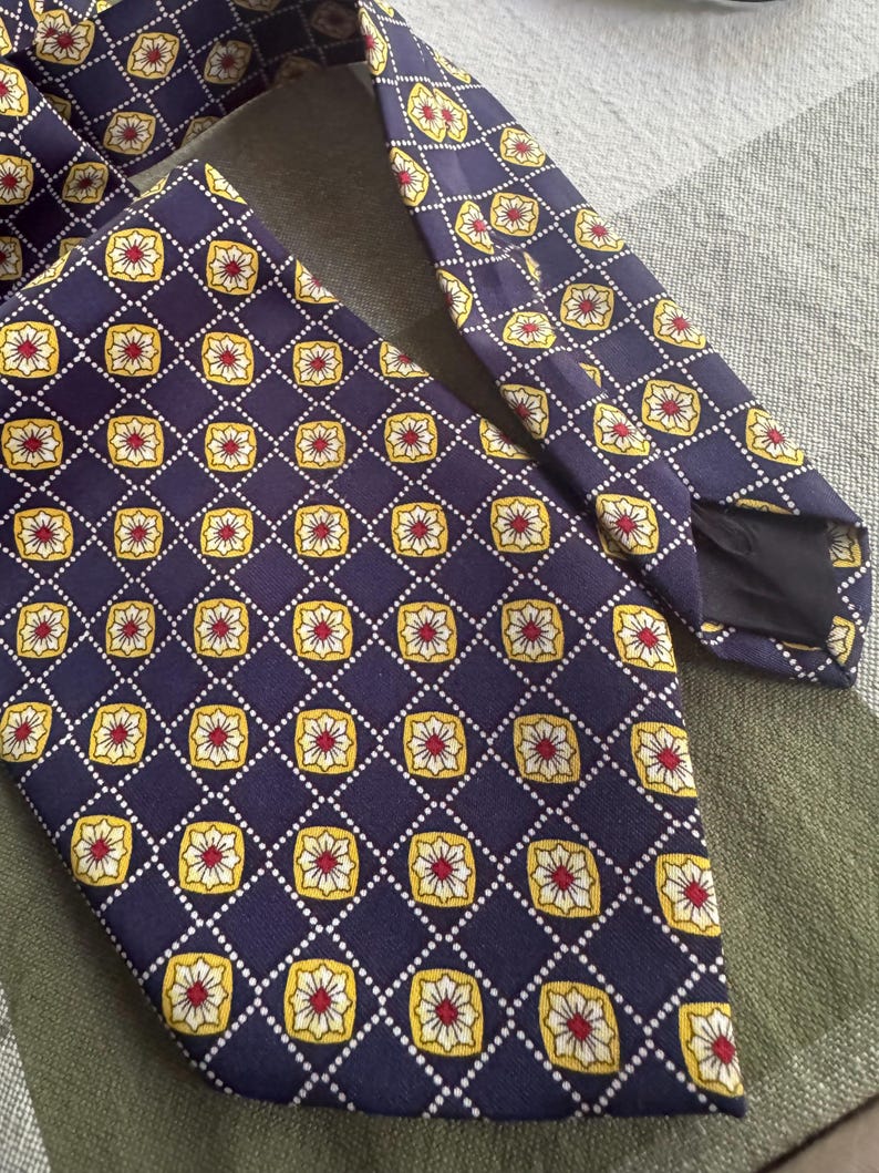 Puede incluir: Una corbata de color morado oscuro con un patr&oacute;n geom&eacute;trico repetitivo de l&iacute;neas de diamantes punteadas en blanco. Dentro de cada diamante hay un cuadrado amarillo de esquinas redondeadas con un motivo floral blanco y rojo.