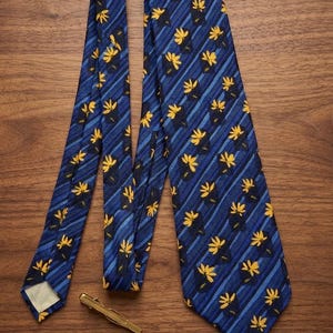 May include: A blue necktie with a repeating pattern of yellow floral designs and diagonal blue stripes. A gold-colored tie clip is also present. The tie is displayed on a wooden surface.