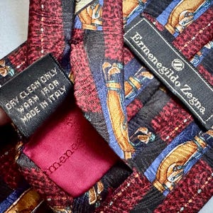 May include: An Ermenegildo Zegna necktie featuring a repeating pattern of golf bags on a background of red, black, and blue plaid. The tie includes a black label with the brand name and a care tag stating Dry Clean Only, Warm Iron, Made in Italy.