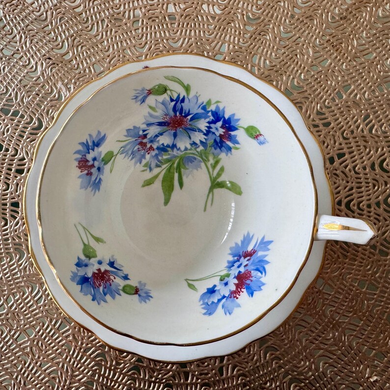 May include: A white teacup with a gold rim and a floral pattern of blue and red flowers. The teacup is on a gold lace doily.