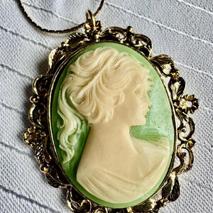 May include: A gold-toned pendant necklace featuring a cameo. The cameo depicts a woman's profile in cream against a light green background, set within an ornate gold-colored frame with floral details. The chain is also gold-toned.