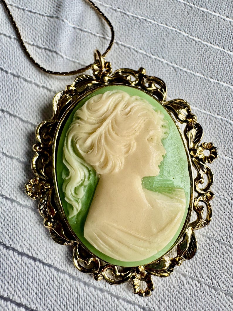 May include: A gold-toned pendant necklace featuring a cameo. The cameo depicts a woman's profile in cream against a light green background, set within an ornate gold-colored frame with floral details. The chain is also gold-toned.