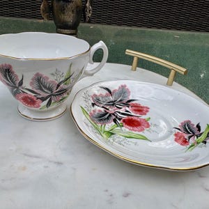 Puede incluir: Un juego de taza y platillo blancos con un dise&ntilde;o floral. La taza y el platillo presentan un patr&oacute;n de flores rosas, negras y verdes con ribete dorado. El juego est&aacute; colocado sobre una superficie de m&aacute;rmol.