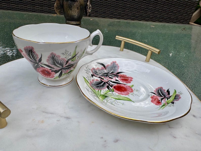 Puede incluir: Un juego de taza y platillo blancos con un dise&ntilde;o floral. La taza y el platillo presentan un patr&oacute;n de flores rosas, negras y verdes con ribete dorado. El juego est&aacute; colocado sobre una superficie de m&aacute;rmol.