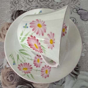 May include: White teacup and saucer set with a hand-painted floral design. The cup and saucer feature pink daisies with yellow centers and green stems. The set has a delicate, vintage aesthetic.