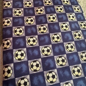 May include: A necktie featuring a repeating pattern of black and white soccer balls set against a checkered background of alternating dark blue and light blue squares. The fabric has a textured appearance with a consistent grid layout throughout.