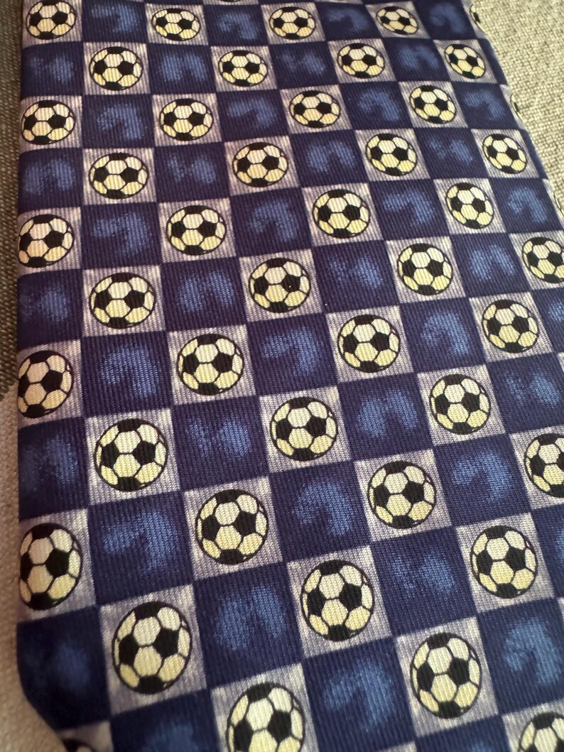 May include: A necktie featuring a repeating pattern of black and white soccer balls set against a checkered background of alternating dark blue and light blue squares. The fabric has a textured appearance with a consistent grid layout throughout.