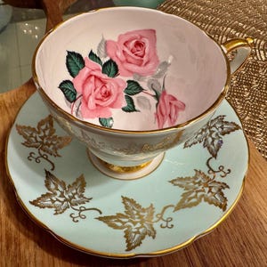 May include: A vintage teacup and saucer set with a delicate floral design. The teacup features pink roses and green leaves on a white background. The saucer is a pale blue colour with gold leaf accents.