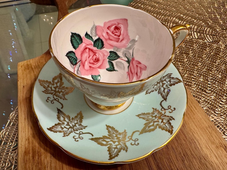 May include: A vintage teacup and saucer set with a delicate floral design. The teacup features pink roses and green leaves on a white background. The saucer is a light blue color with gold leaf accents.