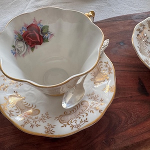 May include: A white teacup and saucer with a gold floral design and a red rose. The teacup has a scalloped edge and a gold handle.
