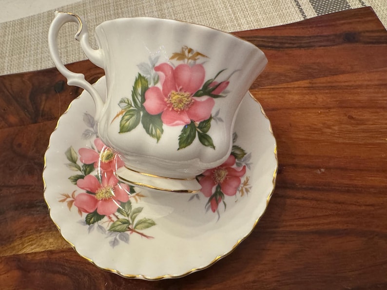 Puede incluir: Una taza de t&eacute; y un platillo blancos con un borde dorado y un patr&oacute;n floral con rosas rosas.