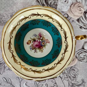 May include: A vintage teacup and saucer set. The teacup features a cream-colored exterior with gold trim and a teal interior with a floral design. The saucer has a matching cream and gold rim. The teacup handle is gold.