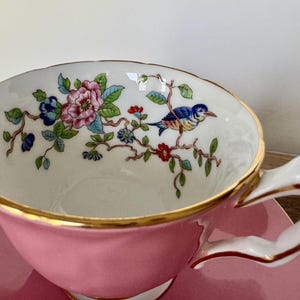 May include: Pink teacup and saucer set with gold trim. The teacup interior features a floral and bird design in blue, pink, and green. The handle is white with gold accents. The saucer is pink.