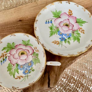 May include: A set of two white porcelain teacups and saucers with a floral design. The cups and saucers have a gold trim and feature pink and blue flowers with green leaves.