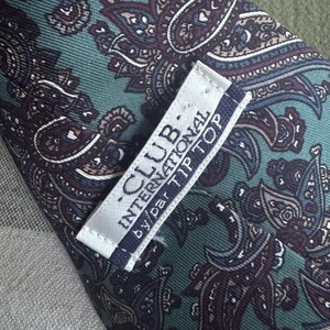 K&ouml;nnte beinhalten: Eine Krawatte mit t&uuml;rkisfarbenem Hintergrund und einem dunkelvioletten sowie schwarzen Paisley-Muster. Auf der R&uuml;ckseite ist ein wei&szlig;es Stoffetikett mit dem Text CLUB INTERNATIONAL by par TIP TOP in dunkelblauer und schwarzer Schrift.