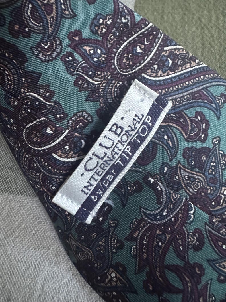 K&ouml;nnte beinhalten: Eine Krawatte mit t&uuml;rkisfarbenem Hintergrund und einem dunkelvioletten sowie schwarzen Paisley-Muster. Auf der R&uuml;ckseite ist ein wei&szlig;es Stoffetikett mit dem Text CLUB INTERNATIONAL by par TIP TOP in dunkelblauer und schwarzer Schrift.