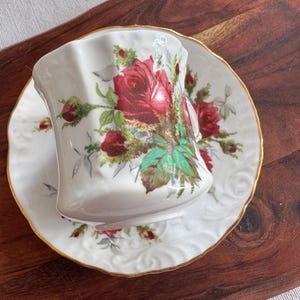 May include: A white teacup and saucer with a gold rim. The cup and saucer are decorated with a floral pattern of red roses and green leaves.