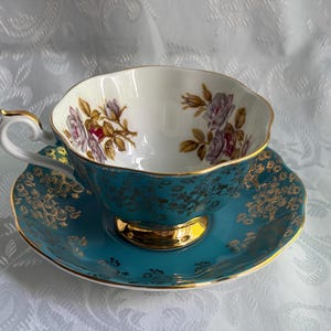 Może przedstawiać: Zestaw filiżanki do herbaty ze spodkiem z turkusowej porcelany, ozdobiony złotym wzorem filigranowym na zewnątrz oraz delikatnym motywem kwiatowym z fioletowymi r&oacute;żami wewnątrz. Filiżanka ma falisty brzeg oraz złoconą podstawę, uszko i krawędź.