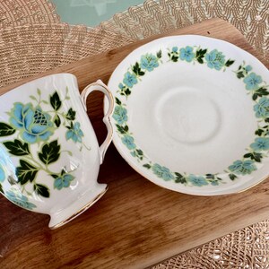 May include: A white teacup and saucer with a floral pattern in shades of blue and green. The floral design features stylised leaves and flowers.