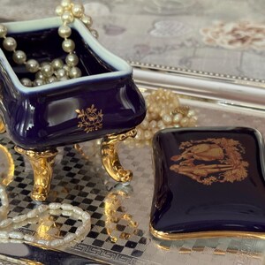 May include: A navy blue porcelain jewelry box with gold accents. The box is in the shape of a small bathtub, resting on gold feet, and contains a pearl necklace. A matching lid sits nearby on a silver tray, with a gold floral design.
