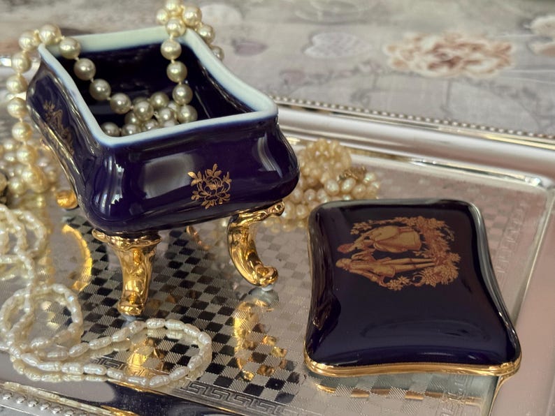 May include: A navy blue porcelain jewelry box with gold accents. The box is in the shape of a small bathtub, resting on gold feet, and contains a pearl necklace. A matching lid sits nearby on a silver tray, with a gold floral design.