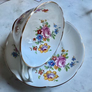 Puede incluir: Juego de taza y platillo blancos con dise&ntilde;o floral. La taza y el platillo tienen un borde festoneado y ribete dorado. El estampado floral incluye rosas rosas, flores azules y amarillas y hojas verdes.