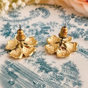 May include: A pair of gold-colored flower-shaped stud earrings. The earrings have a textured surface and a post backing. The earrings are displayed on a white and blue floral patterned plate.