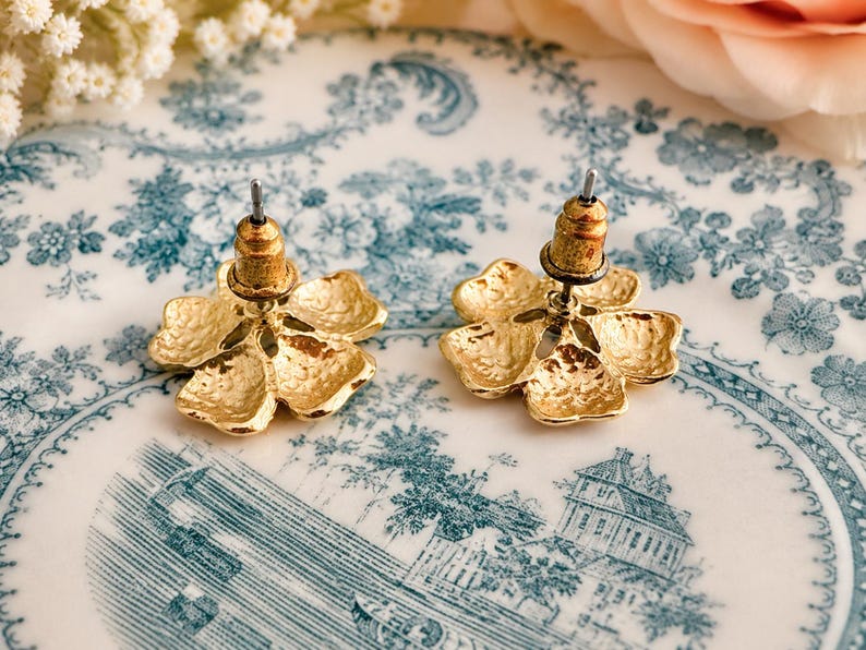 May include: A pair of gold-colored flower-shaped stud earrings. The earrings have a textured surface and a post backing. The earrings are displayed on a white and blue floral patterned plate.