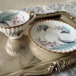 Puede incluir: Un juego de taza y platillo blancos con borde dorado y dise&ntilde;o de helechos y flores. La taza tiene asa y borde festoneado. El platillo es ovalado y tiene un dise&ntilde;o a juego. El juego est&aacute; sobre una bandeja plateada.