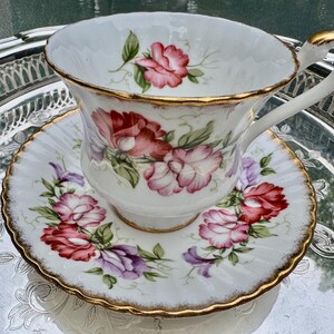May include: White teacup and saucer set with a floral design in shades of red, pink, and purple. The edges are trimmed with gold. The teacup is on a silver tray.