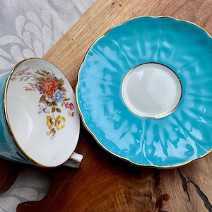 May include: A turquoise teacup and saucer set with a floral pattern. The saucer has a scalloped edge and a gold trim.