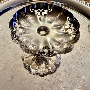 May include: A silver-colored, tiered serving dish with a floral design. The top dish has a scalloped edge and openwork cutouts. The base and tray have an embossed floral pattern. The dish is reflective and sits on a larger tray.