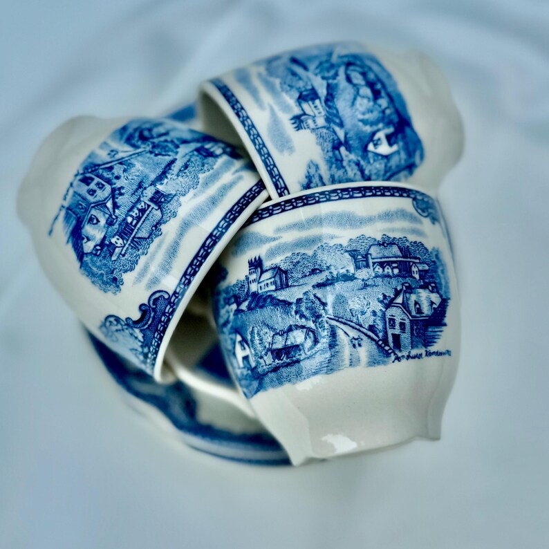 Puede incluir: Cuatro tazas de cer&aacute;mica blanca apiladas, con escenas de paisaje en azul. Las ilustraciones azules y blancas detallan edificios, &aacute;rboles y figuras. Las tazas tienen una est&eacute;tica cl&aacute;sica y vintage, evocando la cer&aacute;mica tradicional.