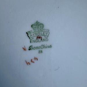 May include: Close-up of a white porcelain plate with a green and gold emblem. The emblem reads "AYNSLEY ENGLAND" with a crown above. The words "Bone China" and the number "29" are also visible. Red numbers are hand-written on the plate.