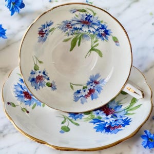 May include: A vintage teacup and saucer set with a delicate floral pattern. The set features a white background with blue and red flowers, accented with gold trim.