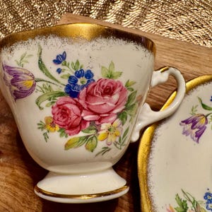 May include: A white teacup and saucer with a gold rim and floral pattern. The flowers are pink, purple, yellow, and blue.