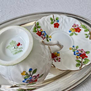K&ouml;nnte beinhalten: Wei&szlig;es Porzellan-Teeservice mit floralem Muster in Rot, Blau, Gelb und Rosa. Die Tasse und Untertasse stehen auf einem silbernen Tablett. Die Tasse hat einen Henkel und steht neben der Untertasse.