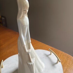 K&ouml;nnte beinhalten: Eine wei&szlig;e Porzellanfigur einer Frau in einem langen Kleid, die einen Regenschirm h&auml;lt. Die Figur steht auf einem wei&szlig;en Marmortablett mit goldenen Griffen. Die Frau hat lange Haare und einen ruhigen Gesichtsausdruck.