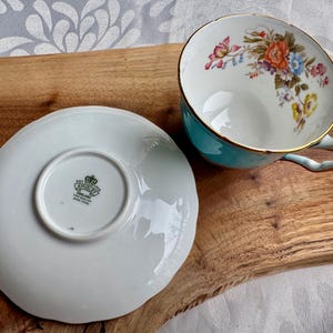May include: A white teacup and saucer with a floral design. The teacup has a turquoise rim and a gold trim. The saucer has a scalloped edge. The teacup and saucer are made by Aynsley England.