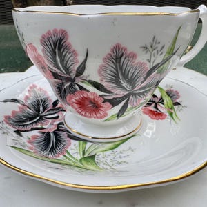 Puede incluir: Juego de taza y platillo blancos con un diseño floral con flores rosas y negras y hojas verdes. Los bordes están adornados con dorado. La taza tiene un asa y se apoya en un platillo a juego.