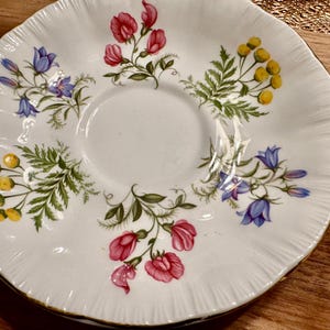 May include: White porcelain saucer with a floral design featuring pink, blue, and yellow flowers. The saucer has a scalloped edge and a gold trim.