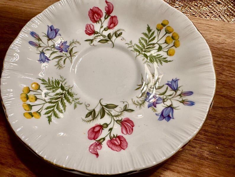 May include: White porcelain saucer with a floral design featuring pink, blue, and yellow flowers. The saucer has a scalloped edge and a gold trim.