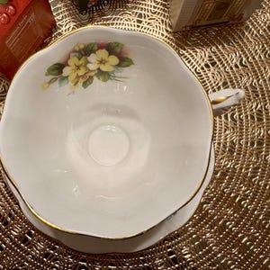 May include: A white teacup and saucer with a floral design featuring yellow and white flowers. The cup has a gold rim and the saucer has a gold trim.
